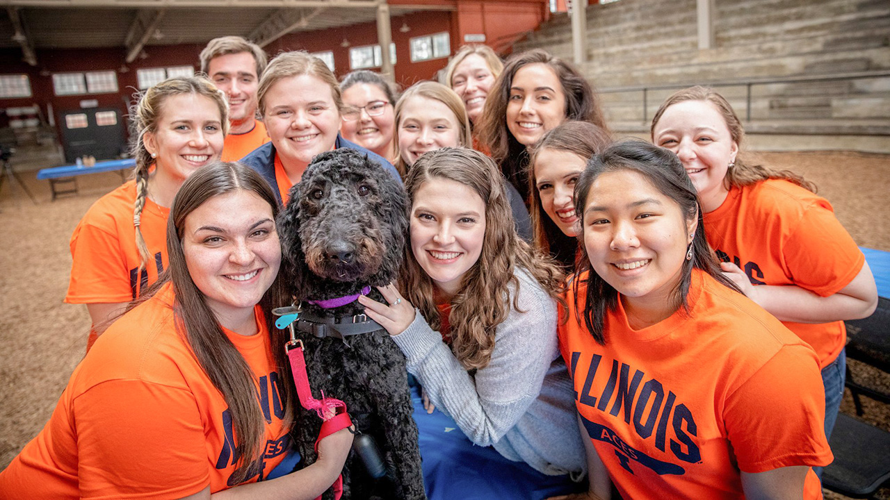 Group of smiling students surrounding a black labradoodle pup.